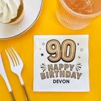 Napkin with '90 Happy Birthday Devon' text on a yellow table with a cake, cup, and fork.