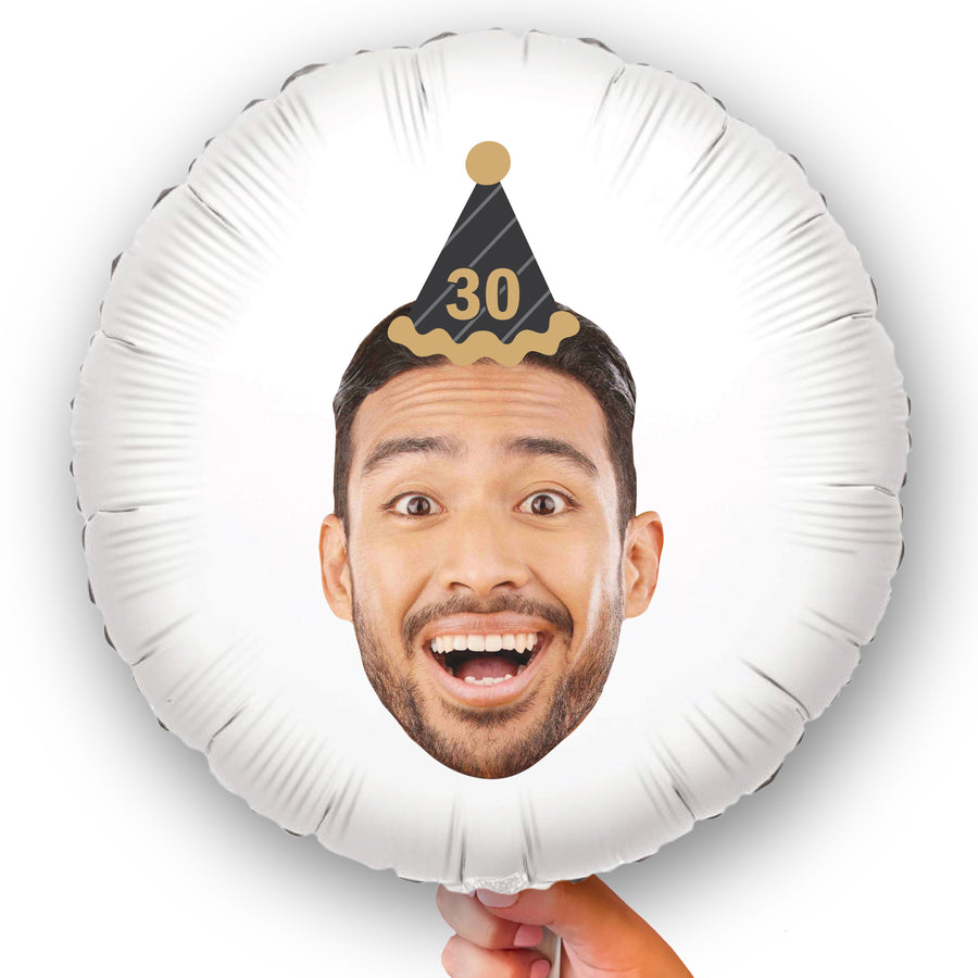 A custom balloon with a photo print of a person's face smiling, with a black and gold party hat on, indicating a birthday celebration.