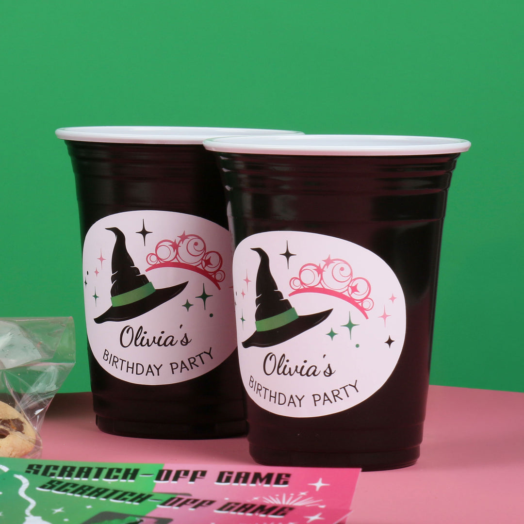 Two custom black party cups with a witch hat design and 'Olivia's Birthday Party' text on a pink surface with a green background.