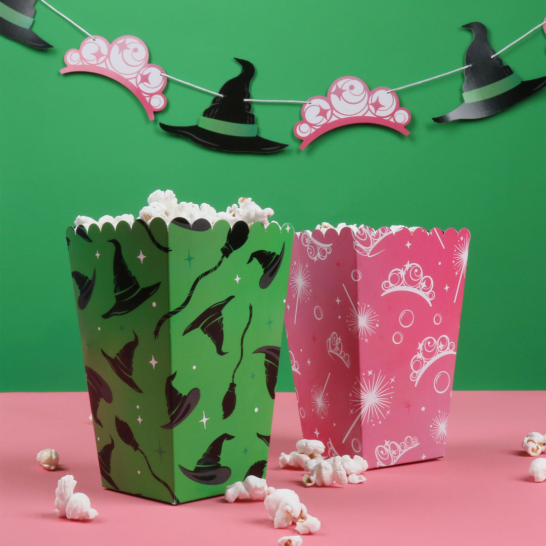 Two popcorn boxes, one green with witch hats and one pink with tiaras, on a pink surface with a green background.