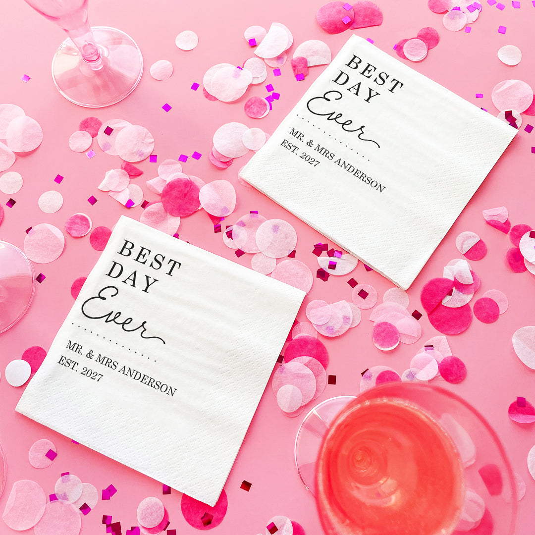 Two 'Best Day Ever' napkins on a pink background with confetti and glasses.