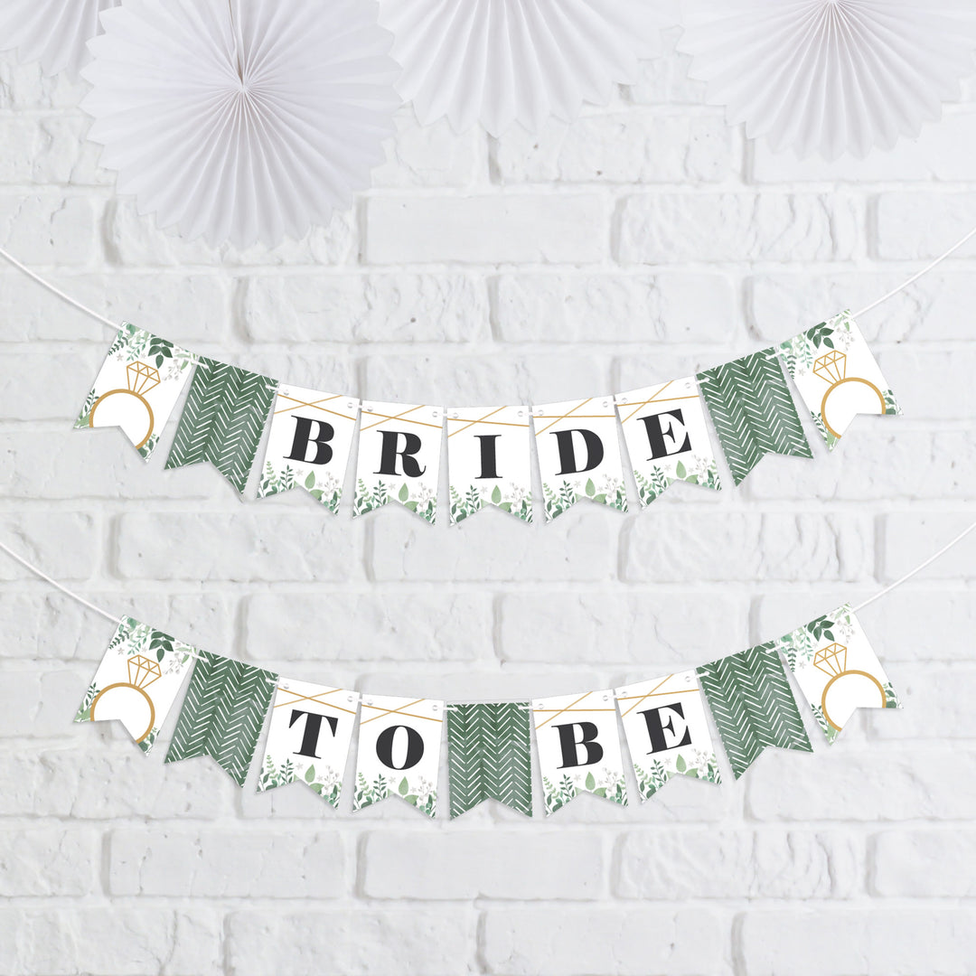 A mini pennant banner with greenery design, spelling out 'BRIDE TO BE' with decorative flags, displayed against a white brick wall.