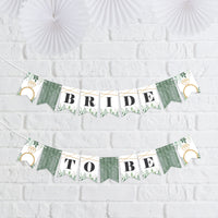 A mini pennant banner with greenery design, spelling out 'BRIDE TO BE' with decorative flags, displayed against a white brick wall.