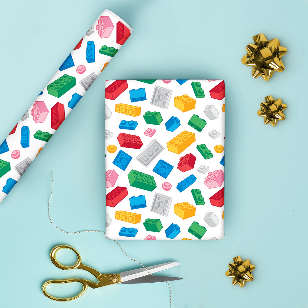 Colorful building block patterned gift wrap paper with scissors and gold bows on a light blue background