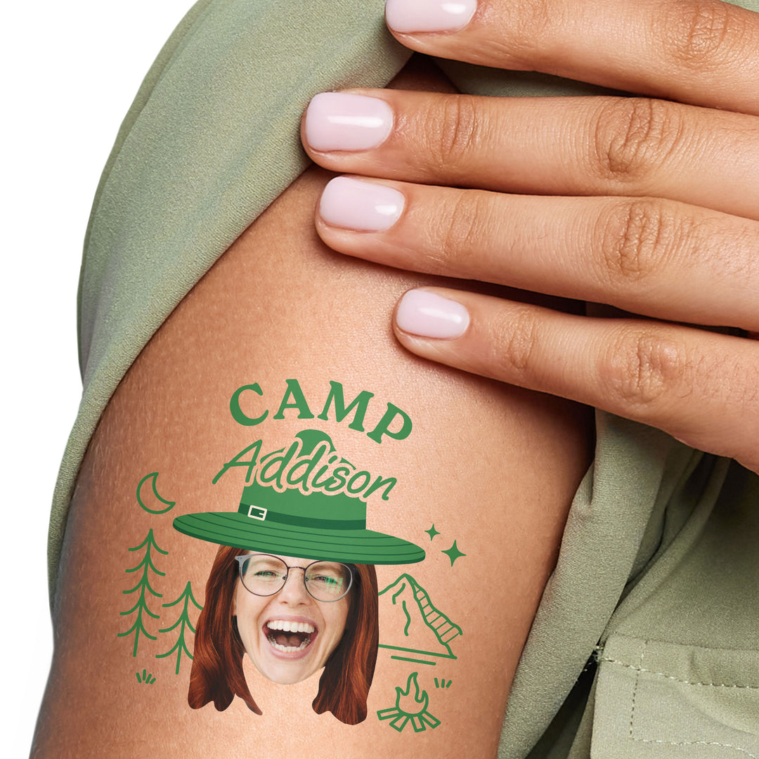 A close-up image of a person's arm featuring a custom face temporary tattoo with the text 'CAMP Addison' and a cartoon hat.
