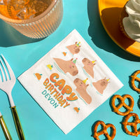 Napkin with cartoon pigs and 'Happy Birthday Devon' text on a blue surface with snacks and cutlery.
