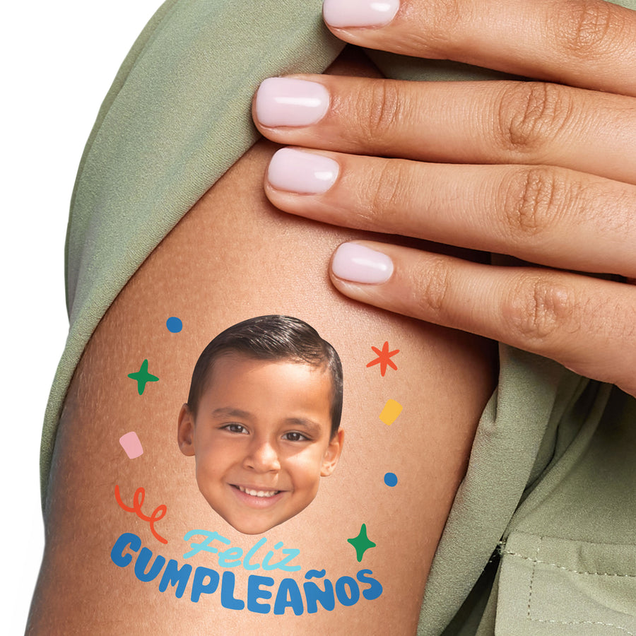 A close-up image of a person's arm with a custom temporary tattoo that has 'Feliz Cumpleaños' written in Spanish with birthday decorations, showing an uploaded photo of a child.
