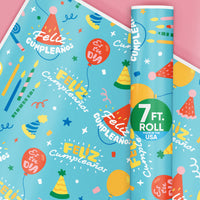 A roll of Feliz Cumpleanos wrapping paper with a fun blue birthday party design, featuring balloons and party hats, wrapped around a pink background.