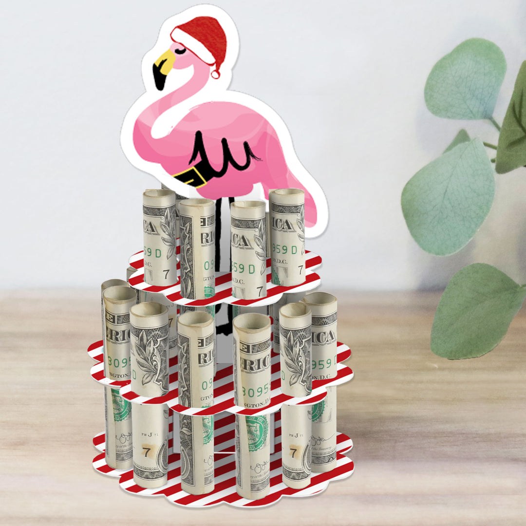 A DIY tropical Christmas party money holder gift featuring a stand shaped like a flamingo with rolled-up bills placed in the slots of a 2-tier cake. The stand is decorated with red and white stripes and a pink flamingo is attached at the top.