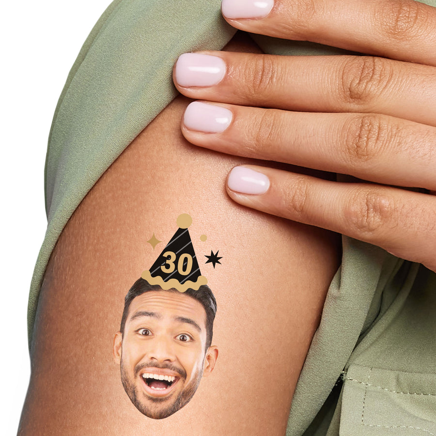A close-up image of a person's arm with a temporary tattoo of a smiling person wearing a black and gold party hat.