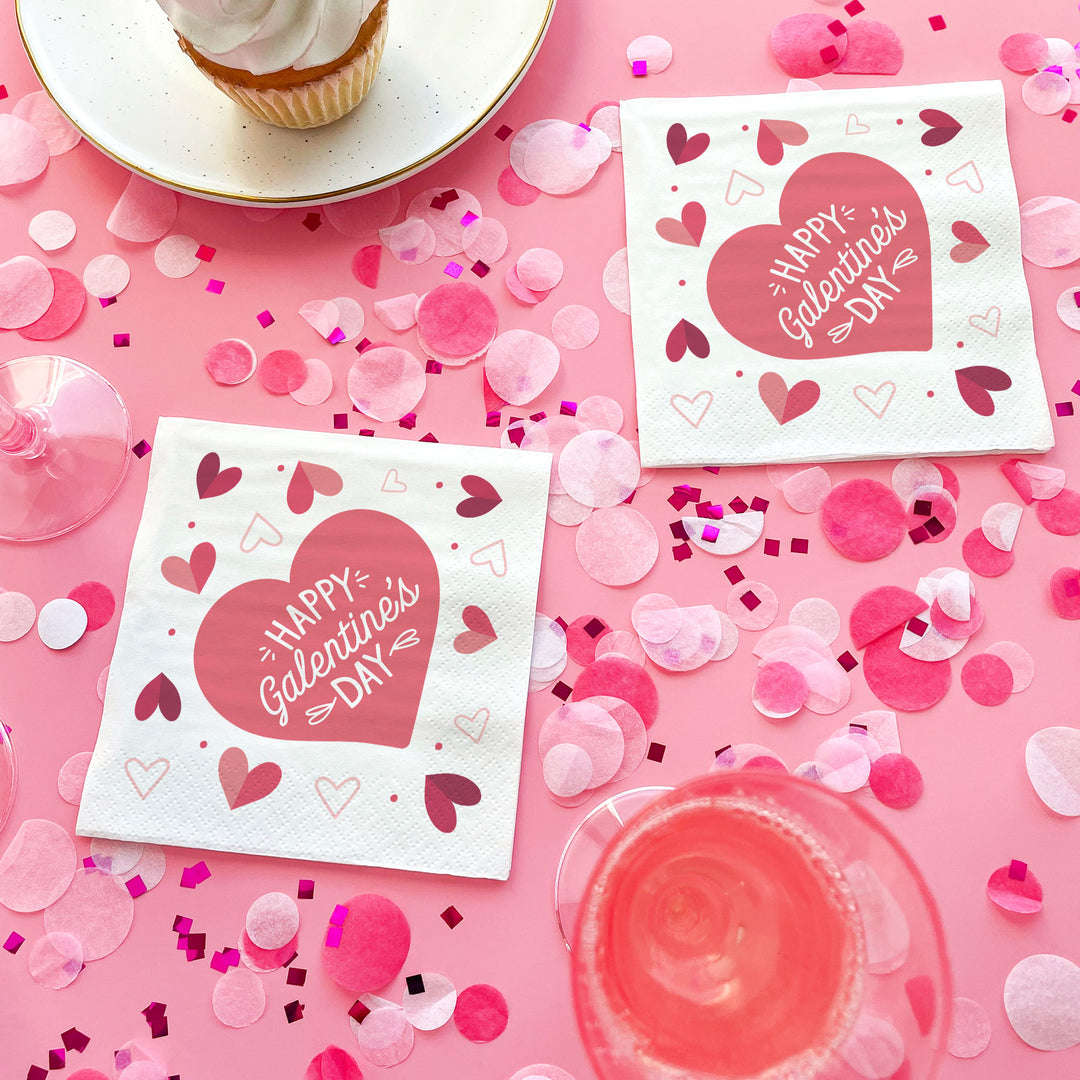 Two 'Happy Galentine's Day' napkins on a pink background with heart confetti and a cupcake.