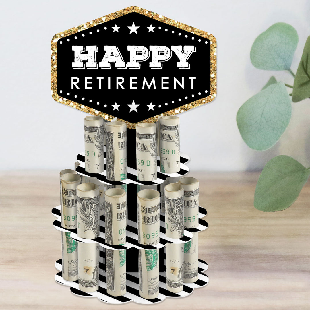 A decorative 'Happy Retirement' themed money holder gift made to look like a two-tier cake with slots for holding rolled-up bills, placed on a wooden surface with a plant in the background.