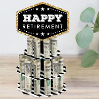 A decorative 'Happy Retirement' themed money holder gift made to look like a two-tier cake with slots for holding rolled-up bills, placed on a wooden surface with a plant in the background.