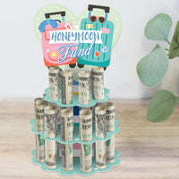 A colorful DIY wedding or engagement party money holder gift designed as a 2-tier cake with 'Honeymoon Fund' text, made from cardboard and holding rolled-up bills in slots.