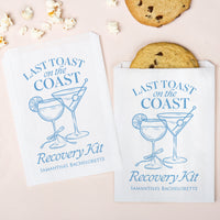 Two 'Last Toast on the Coast Recovery Kit' packages with cookies and popcorn on a light pink background.