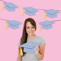 Young woman holding a lavender graduation cap against a pink background with paper graduation caps.