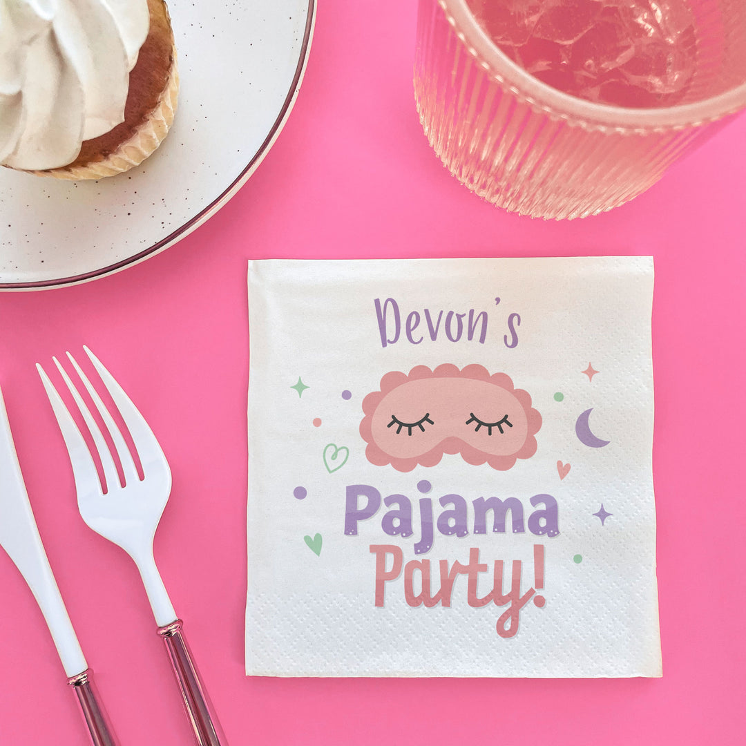 Napkin with 'Devon's Pajama Party!' text on a pink tablecloth with cutlery and dessert.