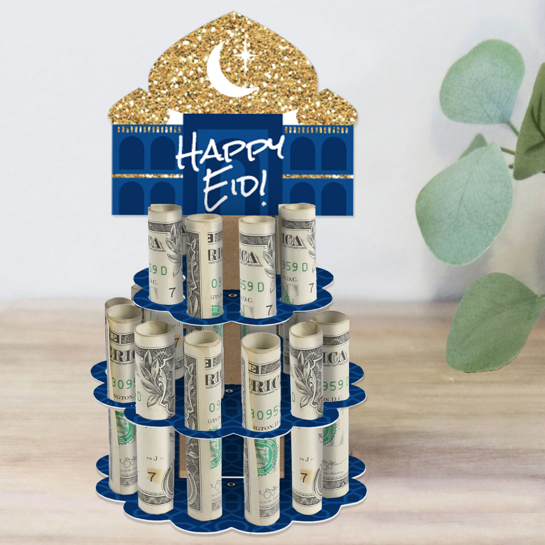 A two-tiered blue and gold-colored paper stand with 'Happy Eid' text on the top, designed to hold rolled-up bills for a party gift, placed on a wooden surface with a green plant in the background.