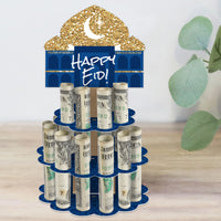 A two-tiered blue and gold-colored paper stand with 'Happy Eid' text on the top, designed to hold rolled-up bills for a party gift, placed on a wooden surface with a green plant in the background.