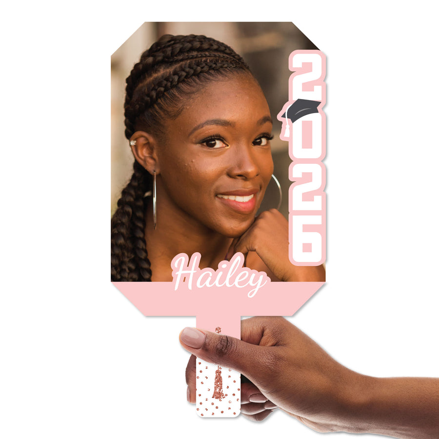 Hand holding a pink custom graduation paddle with a photo of a woman and the name 'Hailey' on a white background