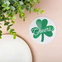 Circular coaster with shamrock design and 'Cheers' text on a beige surface with green leaves.