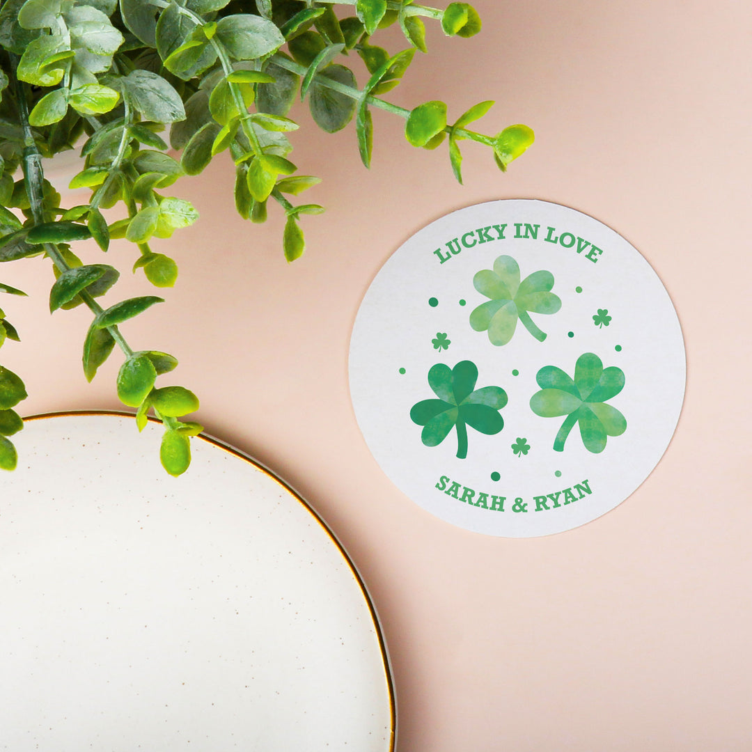 Round drink coaster with green shamrocks and text 'Lucky in Love Sarah & Ryan' on a beige background with a plant.