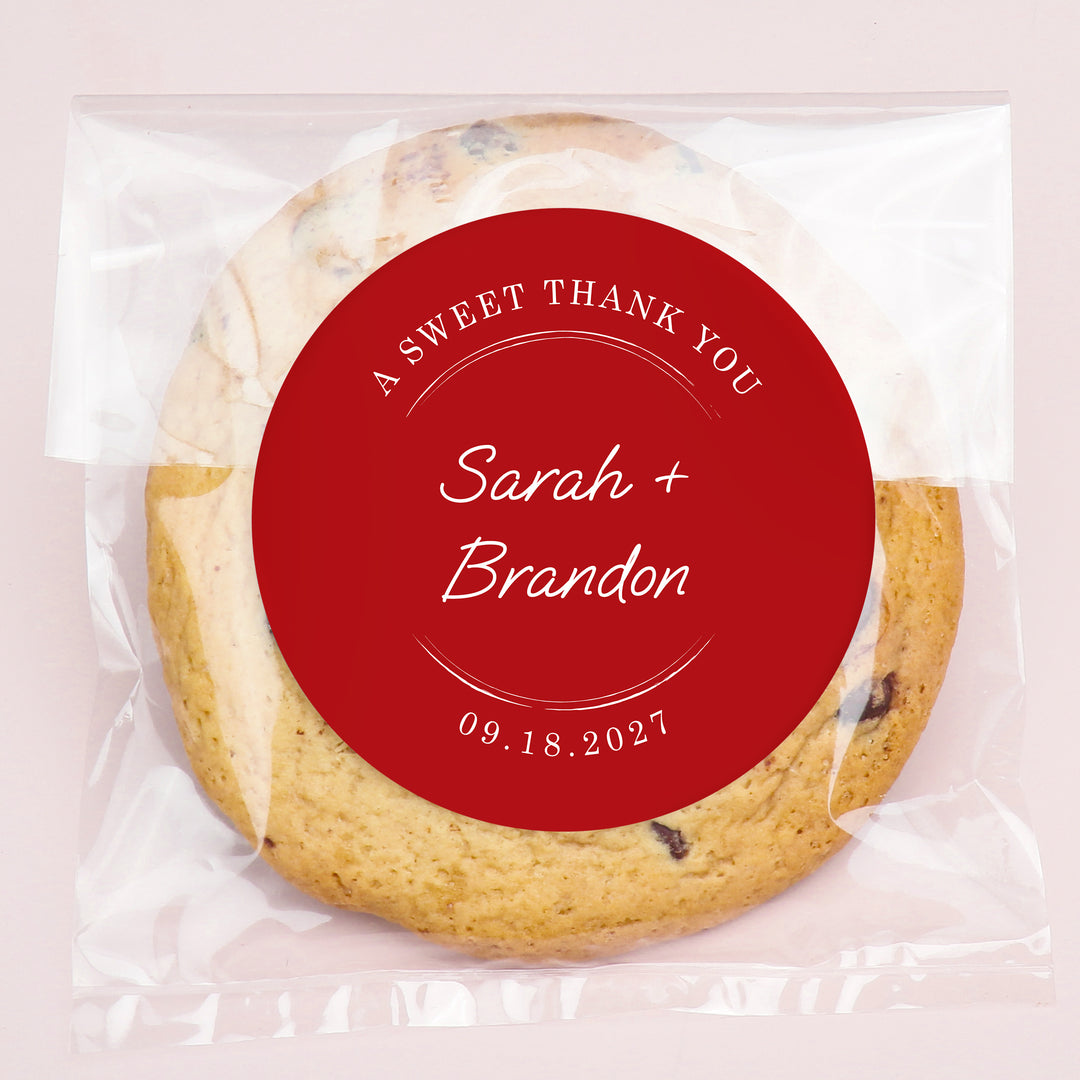 Cookie with a custom red sticker label reading 'A Sweet Thank You Sarah + Brandon 09.18.2027' on a light pink background