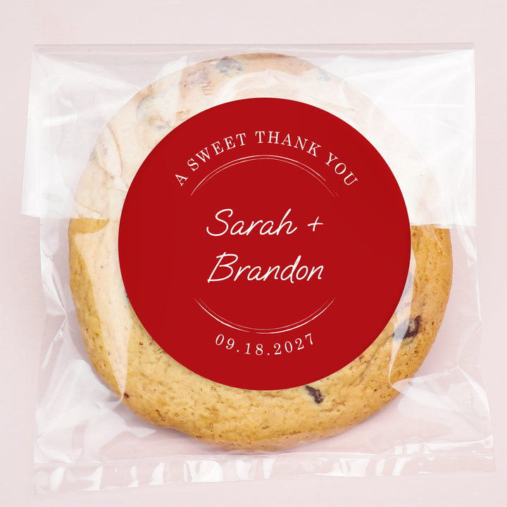 Cookie with a custom red sticker label reading 'A Sweet Thank You Sarah + Brandon 09.18.2027' on a light pink background