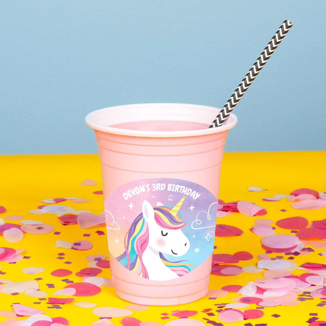 Pink party cup with unicorn design and straw on a yellow surface with confetti