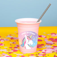Pink party cup with unicorn design and straw on a yellow surface with confetti