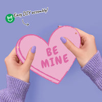 Heart-shaped pink banner with 'BE MINE' text held by hands against a purple background, with 'bg' branding.