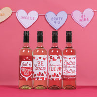 Four bottles of wine with decorative labels on a pink background with heart-shaped signs.