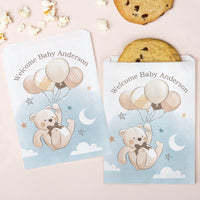 Two baby shower favor bags with teddy bear and balloons design on a pink background with cookies and popcorn.