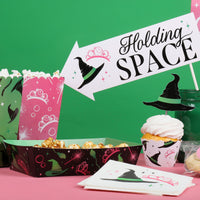 Halloween-themed party setup with popcorn, cupcake, and decorative sign on a pink table against a green background.