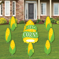 A set of yard signs displayed on a lawn, featuring yellow and green colors with the text 'IT'S CORN' and images of corn ears, indicating a fall harvest theme.