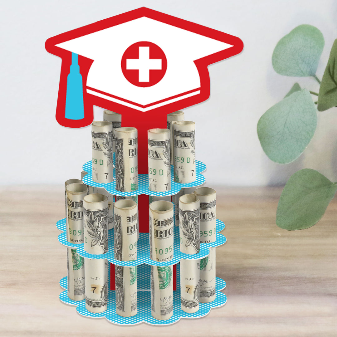 A two-tiered cake-shaped stand made of cardboard designed to hold rolled-up bills, resembling a graduation cash cake with a red grad cap motif on top and a green leaf in the background.