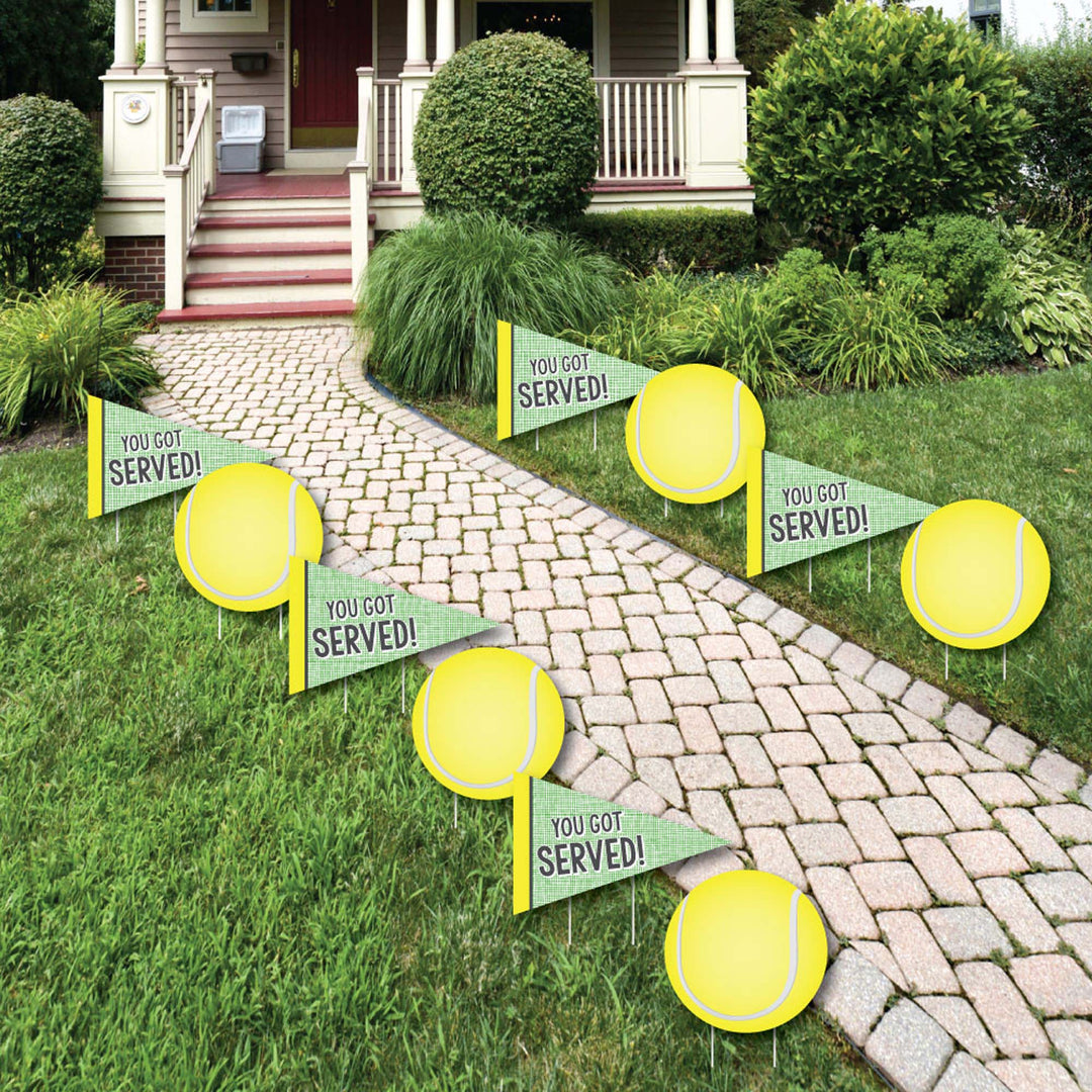 A set of 'You Got Served - Tennis' lawn decorations displayed on a garden path, consisting of yellow tennis ball and flag shaped signs with the text 'YOU GOT SERVED'.