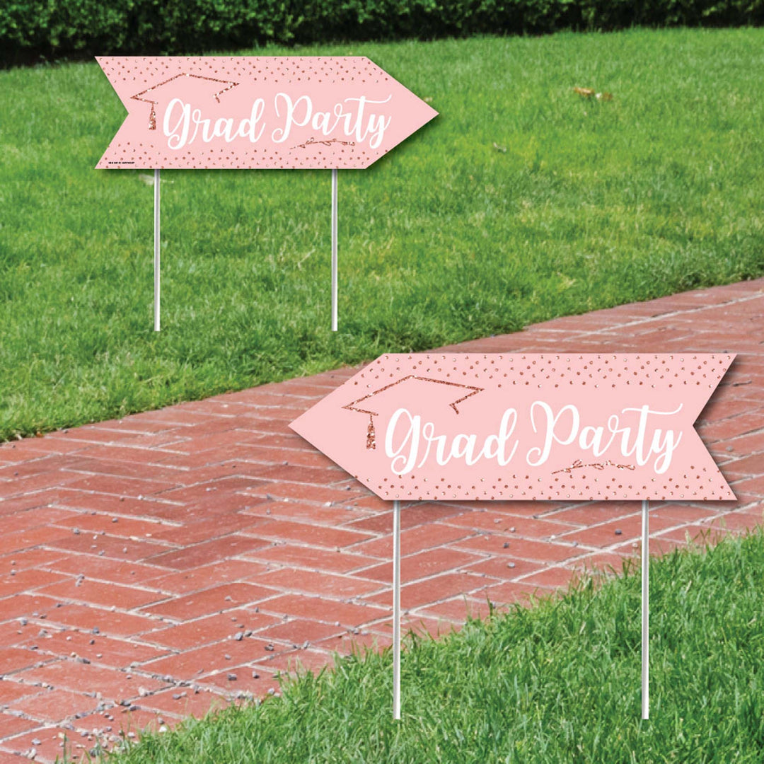 Two rose gold-colored corrugated plastic graduation party directional yard signs with 'Grad Party' text and graduation cap icons, placed on a grassy area beside a brick pathway.
