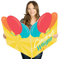 A person holding a large, colorful greeting card with 'Congrats' and 'Woohoo!' text along with balloon illustrations.
