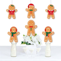 A set of die-cut gingerbread man shaped decorations printed on heavy card stock paper, displayed hanging from strings above a table decorated with white flowers and bottles.
