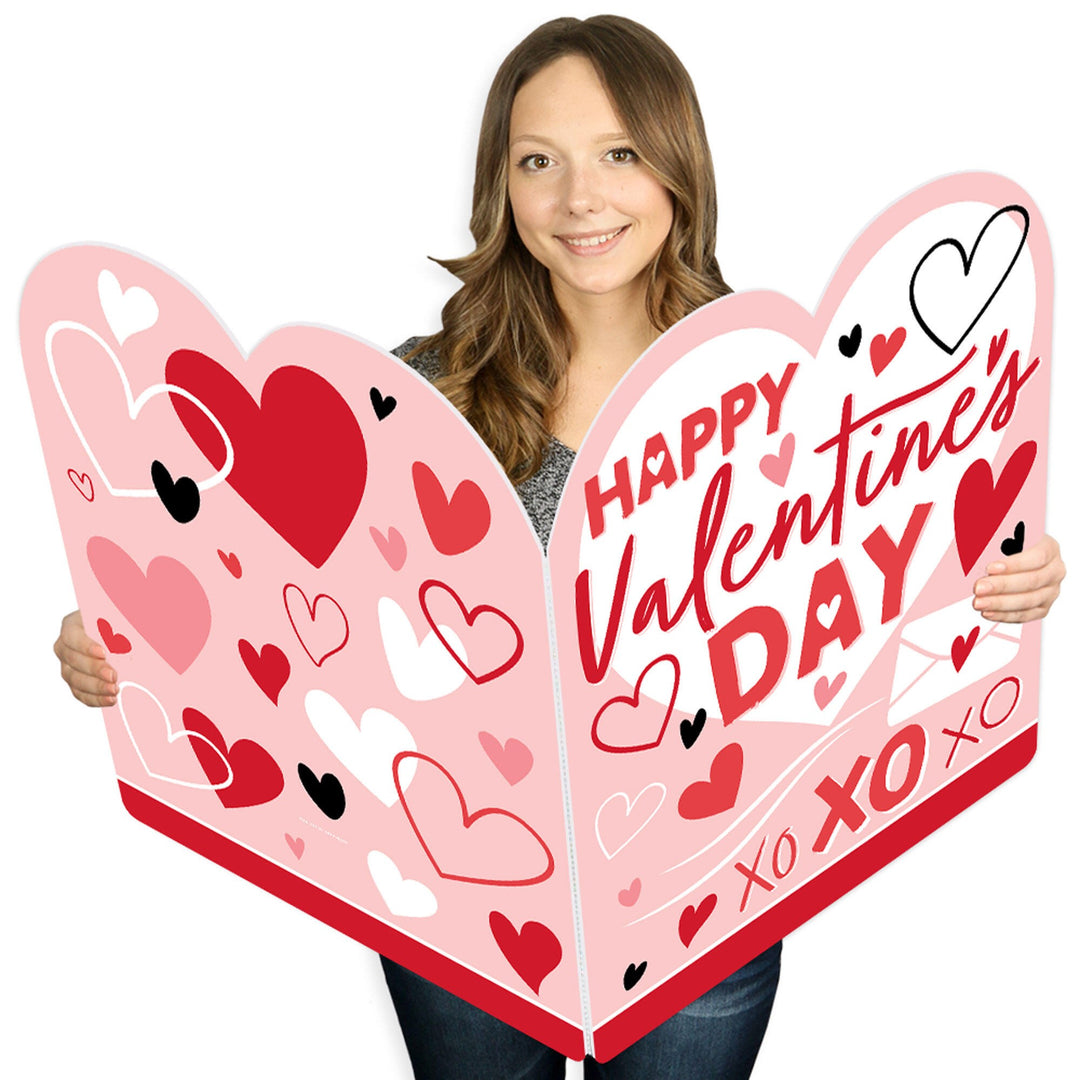 A person holding a large, pink Valentine's Day themed greeting card with text and heart designs