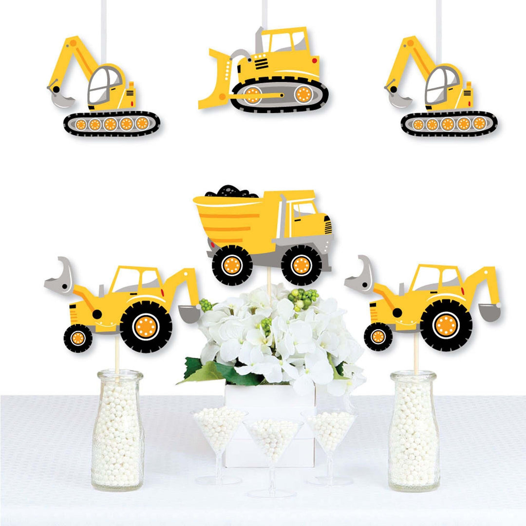A set of construction vehicle shaped party decorations displayed on a table, including cutouts of a dump truck, bulldozer, and excavator in yellow, black, and gray colors.