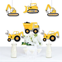 A set of construction vehicle shaped party decorations displayed on a table, including cutouts of a dump truck, bulldozer, and excavator in yellow, black, and gray colors.